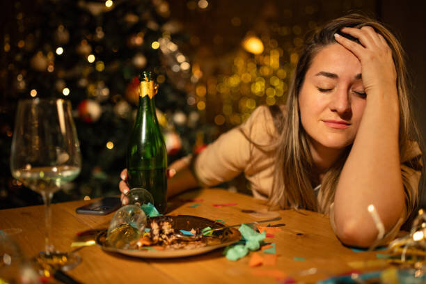 En kvinna sitter vid ett bord med en champagneflaska, tomma glas och konfetti i ett festligt rum, verkar trött eller bekymrad.