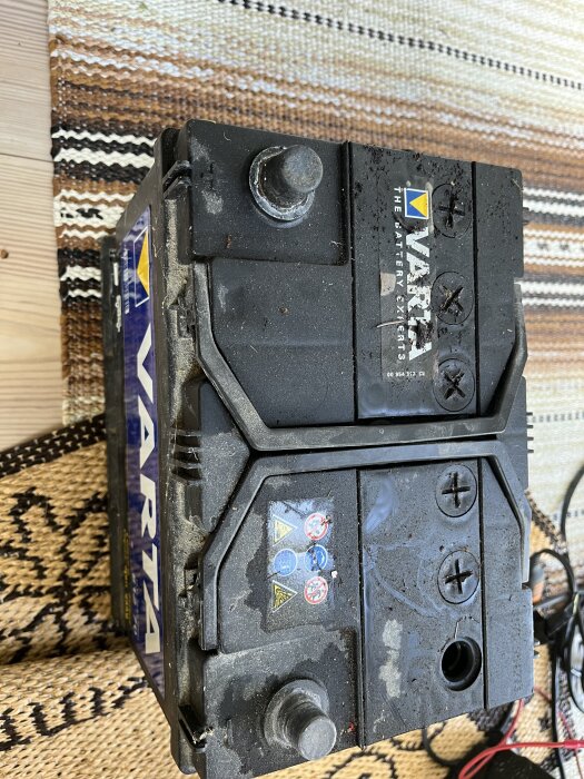 Varta fritidsbatteri med synliga poler och ett öppet hål utan lock, placerat på mönstrad matta.