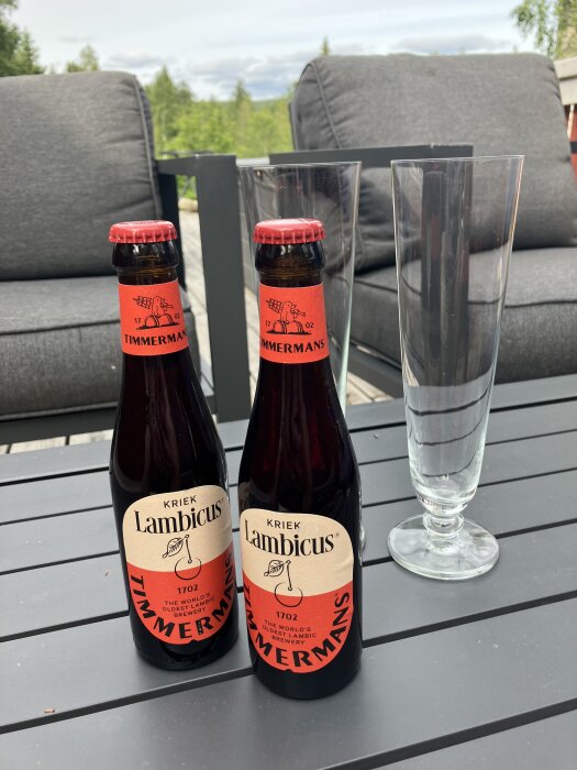 Två flaskor Timmermans Kriek Lambicus öl och två tomma glas på ett utebord med grå soffor i bakgrunden.