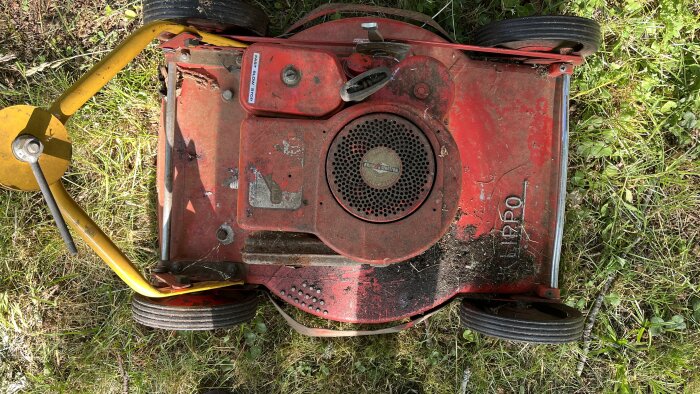 Gammal rödfärgad Klippo gräsklippare med gul handtagsstång, smutsig och använd, fotograferad ovanifrån på gräsmatta.