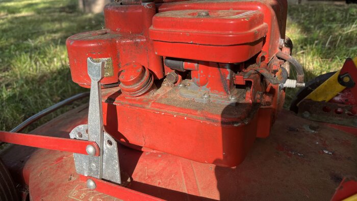 Röd maskindel med metallkomponenter, potentiellt en äldre Klippo gräsklippare sett utomhus med gräs i bakgrunden.