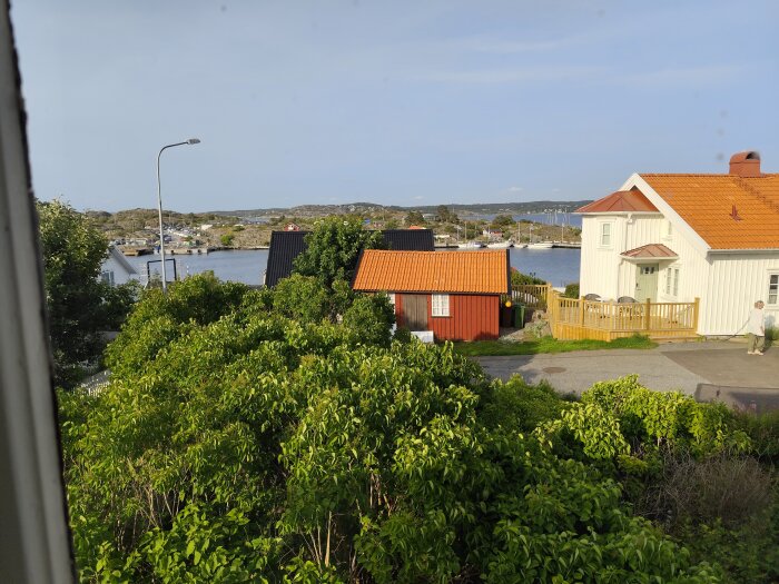 Vy över ett sommarhusområde med trädgårdar, en röd stuga, vita hus och hav i bakgrunden under en klarblå himmel.