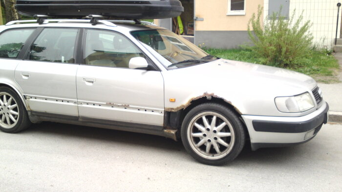 Silverfärgad Audi 100 med rostskador vid hjulhus och biltrösklar, fotograferad på en gata i maj 2013.