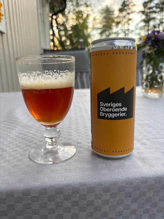 En burk och ett glas med öl från Sveriges Oberoende Bryggerier står på ett bord med vit duk, blommor samt grönska i bakgrunden.