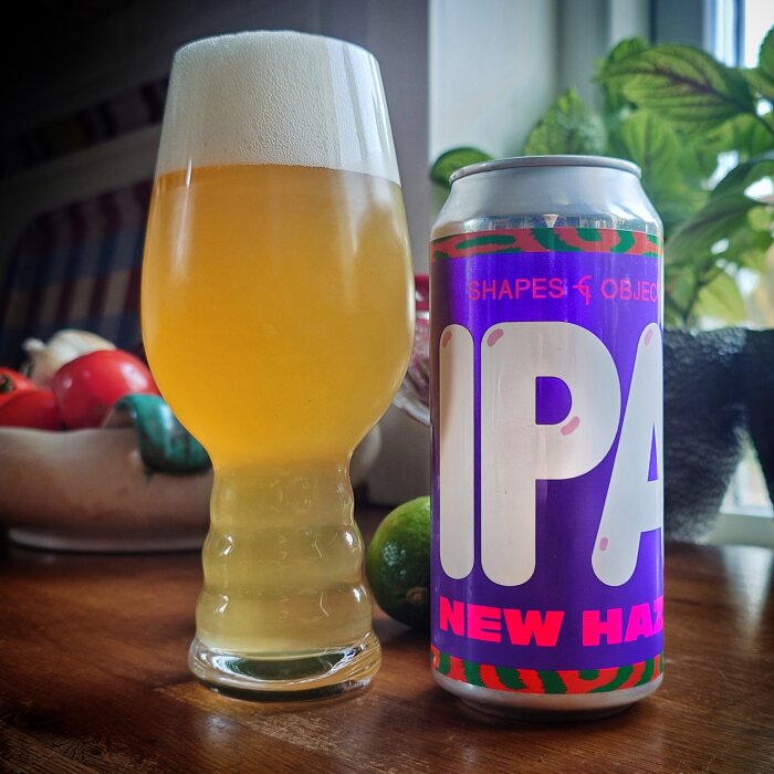 Ett glas med skummande öl bredvid en burk IPA, omgiven av frukt och grönt på ett bord nära ett fönster.