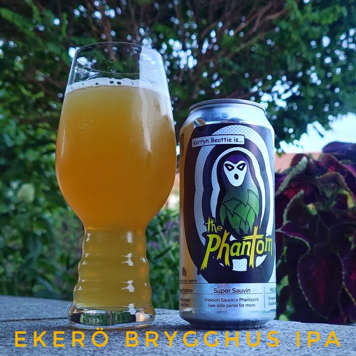 Ett glas fyllt med gul IPA och en burk med etiketten "The Phantom" bredvid, på ett bord utomhus med grönska i bakgrunden.