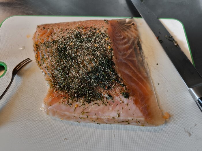 Gravlax på skärbräda med dill och kryddor, en kniv och en gaffel ligger bredvid.