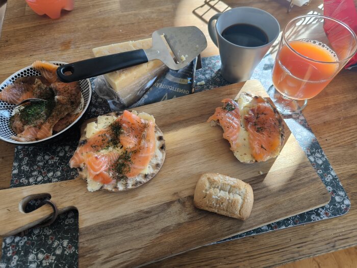 Frukost med gravlax på knäckebröd, Västerbottensost, en kopp svart kaffe, blodgrape juice och en Nyåkers mandelkubb på en träbricka.