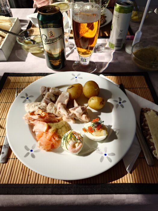 Tallrik med midsommarlunch: inlagd sill, potatis, lax, ägg med topping. Öl och dryck syns i bakgrunden.