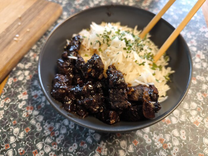 Nötköttsbitar i teriyakimarinad serverade med basmatiris i en skål, dekorerat med örter och ätpinnar på sidan.