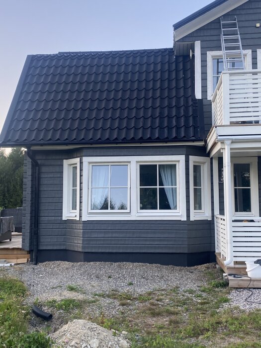 Grått hus med svart tak och vita detaljer, sett från sidan. Grus och lite gräs framför. Stege syns vid sidan av ytterdörren.