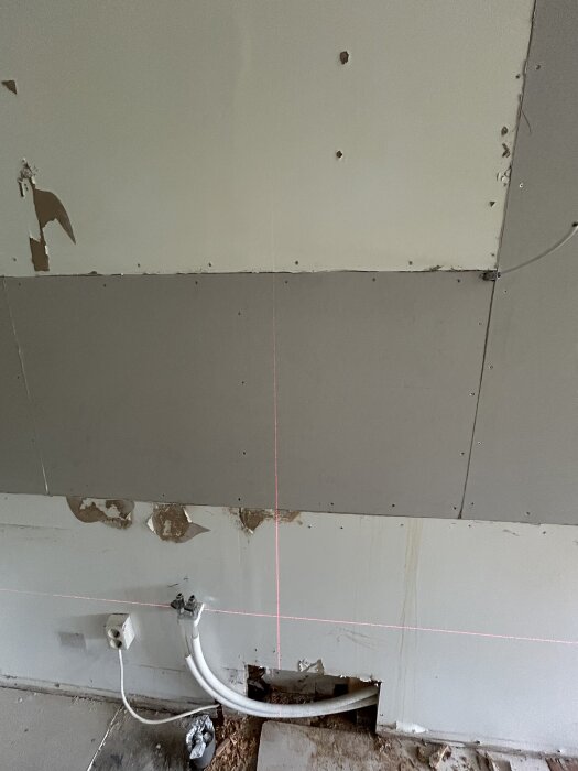 Vägg med gipsplattor där delar av ytan är avskalad, synliga ledningar och laserlinjer markerar vågräta och lodräta punkter för montering.