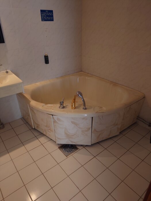 Hörnbadkar i ett kaklat badrum med en vit handfat bredvid. Badkaret har en guldfärgad kran.