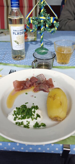 Tallrik med matjessill, brynt smör, gräddfil med gräslök och kokt potatis. Flaskor och glas syns i bakgrunden.