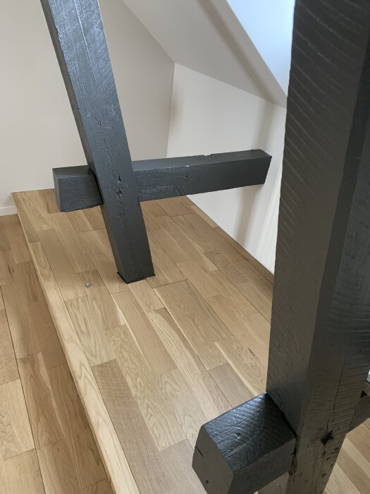 Svartmålade träbjälkar korsar ett golv av ljust trä i ett rum med snedtak.