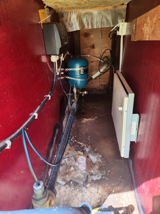 Inuti ett pumphus med ett blått expansionskärl och anslutna ledningar, synlig läcka och vått område på golvet.