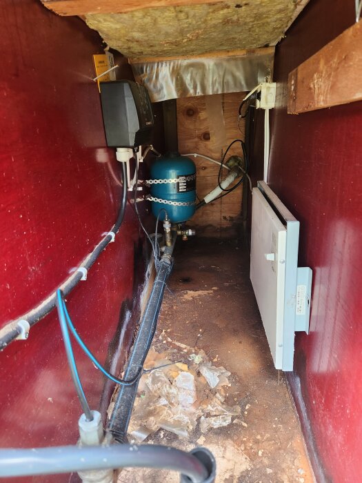 Inuti ett pumphus med en blå vattenpump och tillhörande rör som ligger på ett jordigt golv med lösa materialbitar.