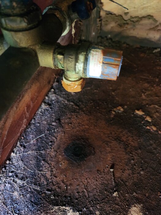 Närbild på en läckande vattenpump med rostiga rörkopplingar, omgivna av smutsigt och delvis fuktigt golv.