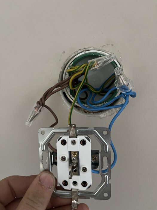 Närbild av ett öppet vägguttag med exponerade elkablar i olika färger anslutna, hålls av en hand i en vägg.