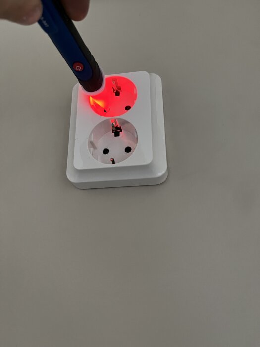 Spänningsdetektorpenna lyser rött vid övre uttaget på ett vägguttag, indikerar ström på ena sidan av uttaget.