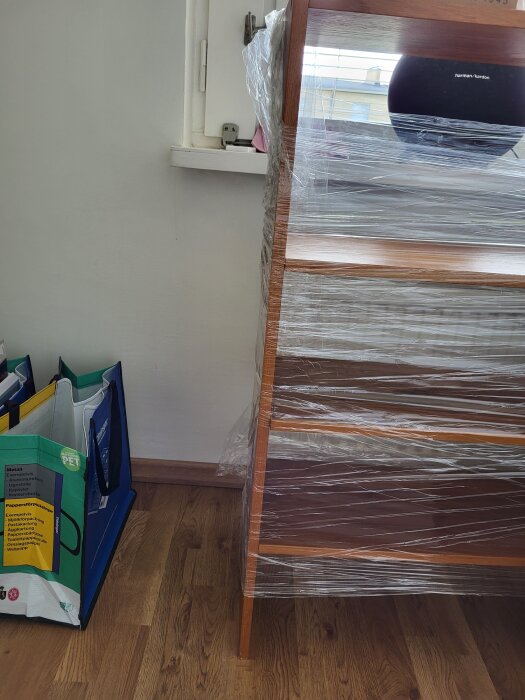 Inplastad gammal bokhylla mot en vit vägg, bredvid står kassar för återvinning. En svart högtalare syns på hyllan.