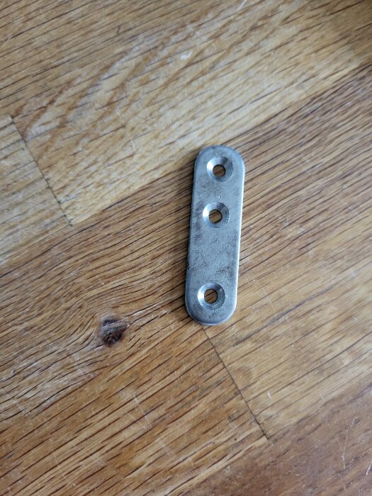 Metallbeslag med tre hål liggande på ett trägolv, används för att fästa ihop bokhylledelar.