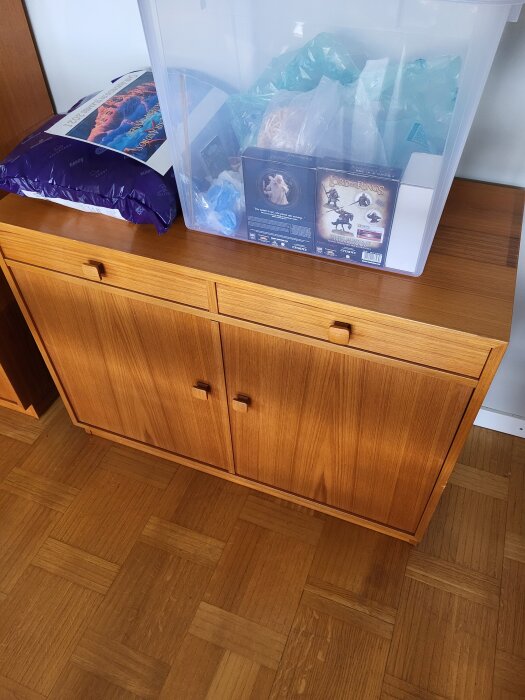 Träskåp med två dörrar och lådor ovanpå. En genomskinlig plastlåda och andra föremål står på skåpet. Parkettgolv ses under skåpet.