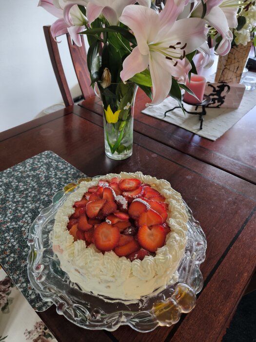 Hemgjord gräddtårta med vispad grädde och skivade jordgubbar, på ett glasfat, står på ett mörkt träbord bredvid en vas med blommor.