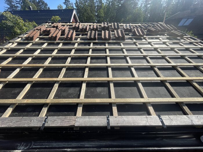 Husets takstruktur med träreglar redo för tegelpannor, några redan på plats, omgiven av grönska och intilliggande hus.