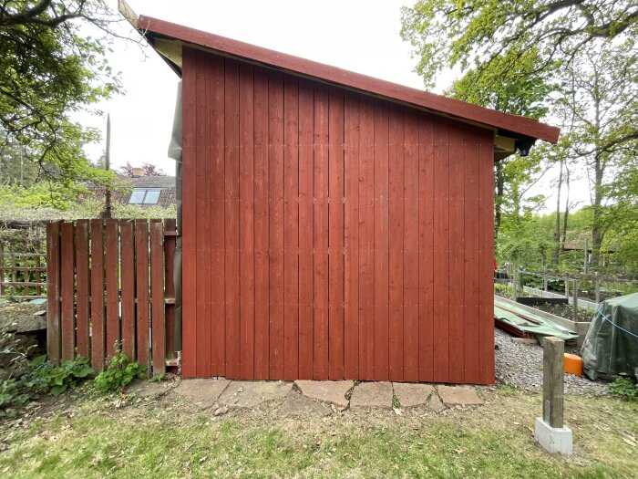 Röd träpanel på en byggnad med luftig trädgård i bakgrunden, omgiven av träd och grönska, samt stenlagd mark framför.