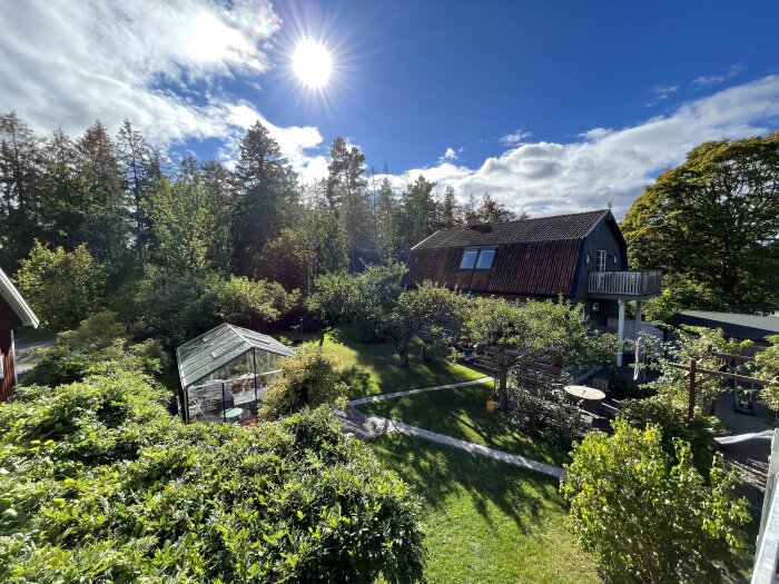 Solig trädgårdsvy med glasväxthus, villa med tegeltak och omgivande träd.