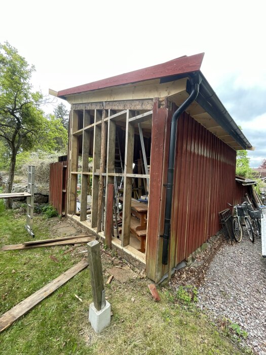 Röd byggnad med delvis borttagen panel, arbetsmaterial framför och cyklar vid sidan, omgiven av träd och stenar.