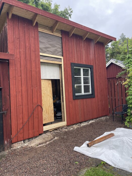 Rödfärgat byggprojekt med nytt fönster installerat i träpanel; en träskiva täcker öppningen för en dörr, grusstig och byggmaterial syns i förgrunden.