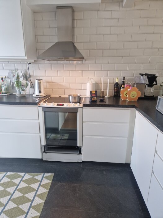 Modernt kök med vita skåp, rostfri fläkt, svart bänkskiva och kaklad vägg. Kokplatta med kastrull syns centralt.