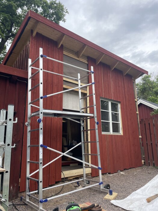Röd byggnad med träpanel, fönster och stege. Pågående renovering, byggnadsställning vid väggen, solitt tak, grusmark framför.