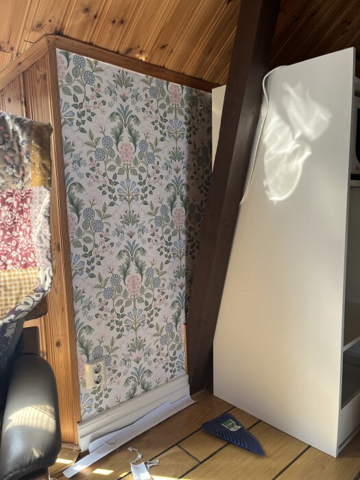 Blommig tapet på vägg, spånskiva lutad mot vägg, träpanel i taket, eluttag nära golvet, material och verktyg på golvet.