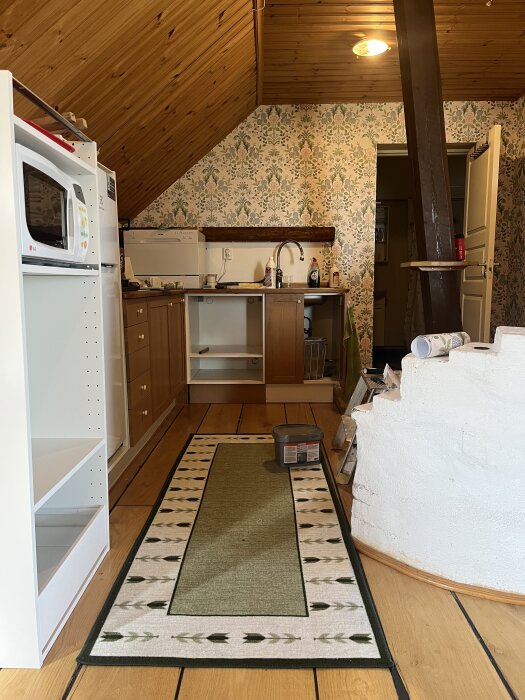 Kök i lägenhet med trägolv och blommig tapet, skåp under bänken, och hyllor. Mikrovågsugn ses till vänster.