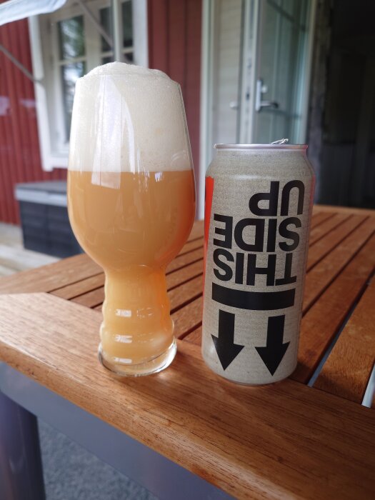 En ölglas fylld med skummande öl bredvid en burk på ett träbord utomhus.