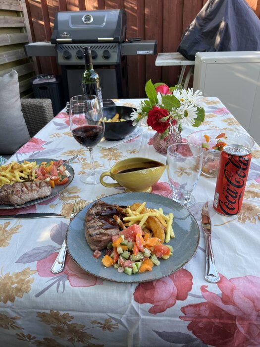 Grillad kalventrecote med pommes, klyftpotatis och sallad på sommardukad uteplats, rödvin och Coca-Cola i glas och burk.