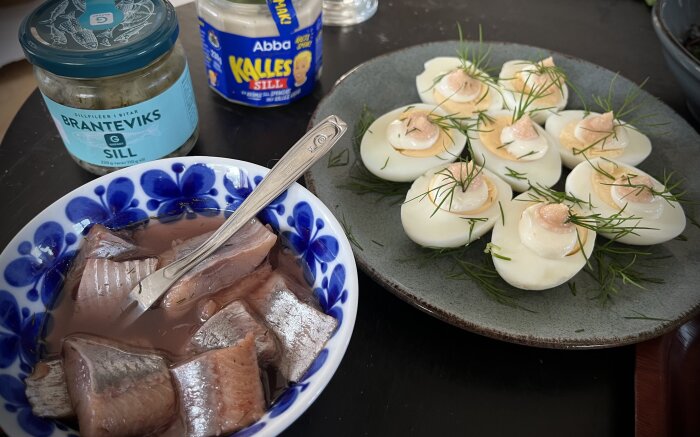 Inlagd sill i skål bredvid ägghalvor med kaviar och dill på fat, med sillburkar i bakgrunden.