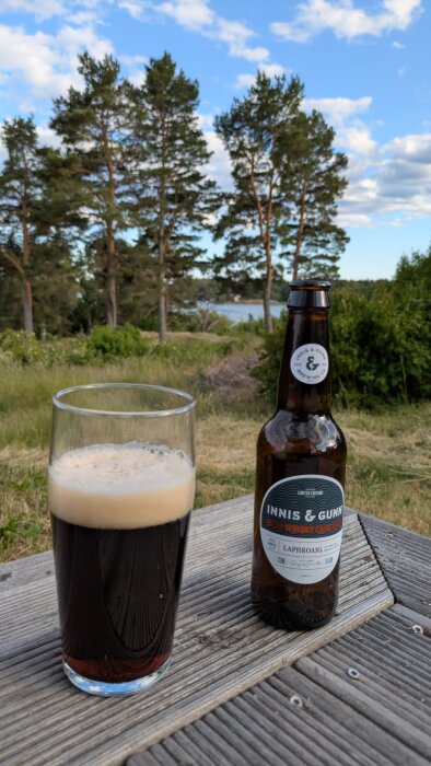 Glas med mörkt öl och en flaska Innis & Gunn Whisky Cask ale på ett bord med tallar och en sjö i bakgrunden under en blå molnig himmel.