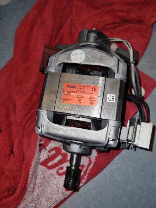 Motor till tvättmaskin liggande på en röd handduk, med etikett som visar märken och tekniska specifikationer.