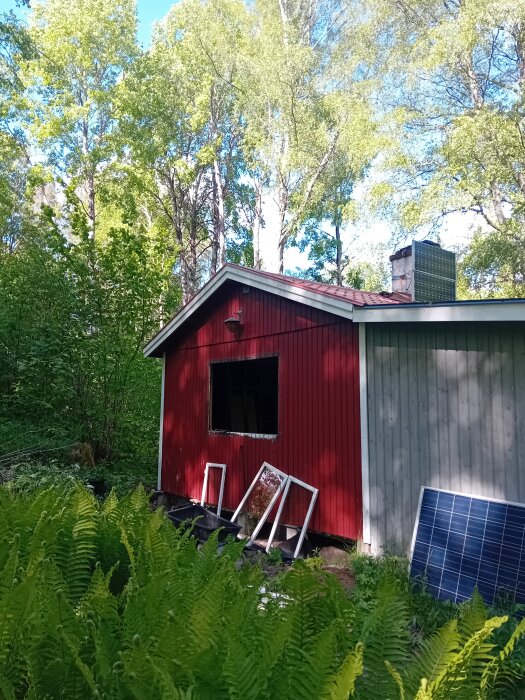 Röd trästuga med ena väggen blågrå, omgiven av skog. Fönsterrutor och en solpanel står lutade mot väggen.