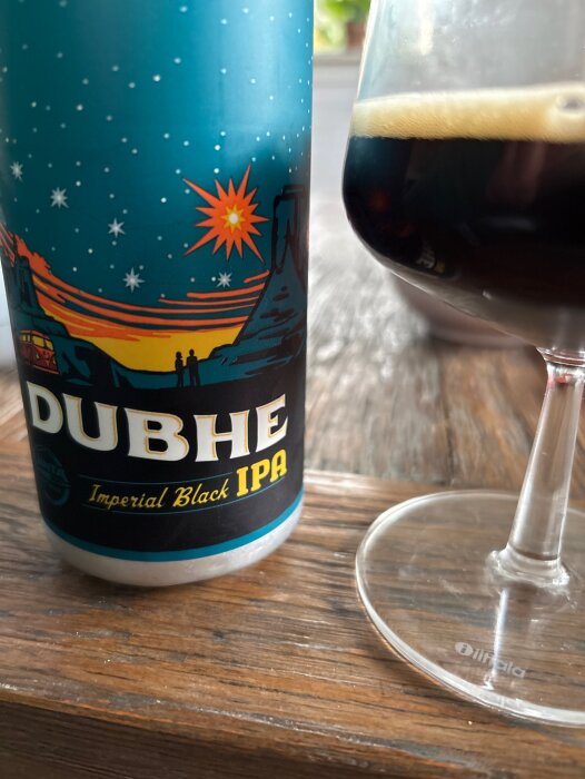 Burk med Dubhe Imperial Black IPA och ett glas med mörk öl på ett träbord.