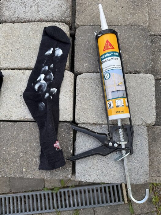 Svart strumpa med vita färgfläckar bredvid en fogpistol med SikaFlex-402 på stenar utomhus.