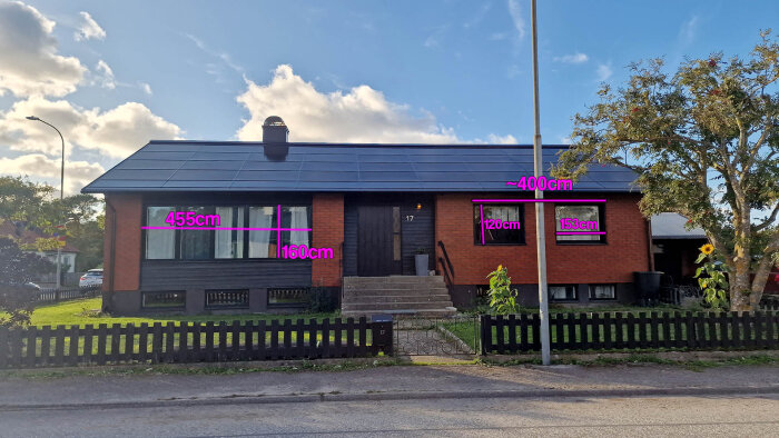 Tegelhus med solpaneler på taket, måttangivelser för fönster och fasad; trädgård med gräsmatta och staket i förgrunden.