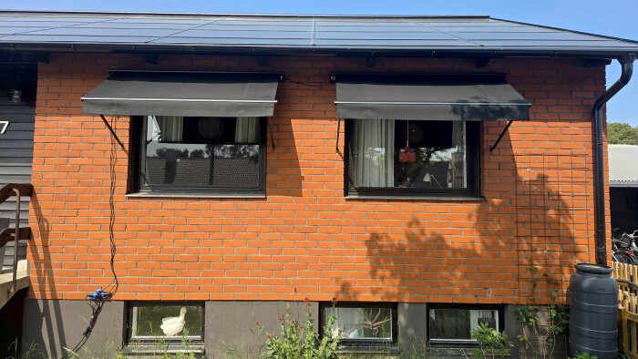 Röd tegelvägg med två fönster och svarta markiser ovanför, del av ett hus.