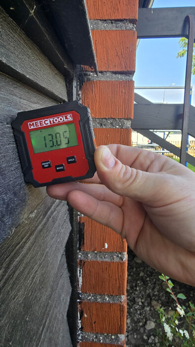 En hand håller en digital vinkelmätare från Meec Tools mot en vägg av svart träpanel och rött tegel, med displayen som visar 13.05 grader.