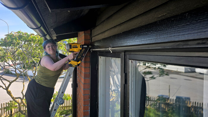 Kvinna på stege använder borrmaskin för att montera konsol vid träfasad och tegelvägg, med träd och staket i bakgrunden.
