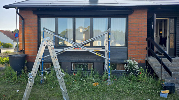 Byggnadsställning framför husfasad med stora fönster och tegelväggar, en stege står lutad intill och trädgårdsarbete pågår runt omkring.